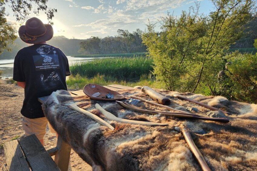 A person standing by a table with tools and a canoe on a sandy beach with a lake and trees in the background. 1