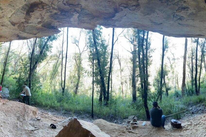 A forest with a large rock formation overhead, a person sitting on the ground, and another person standing nearby. 2