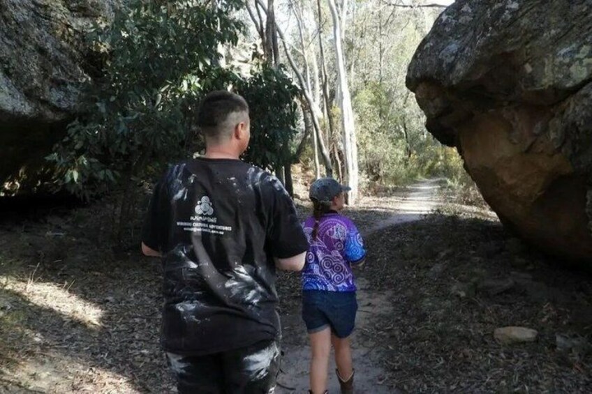Two people walking on a dirt path surrounded by trees and rocks. 3