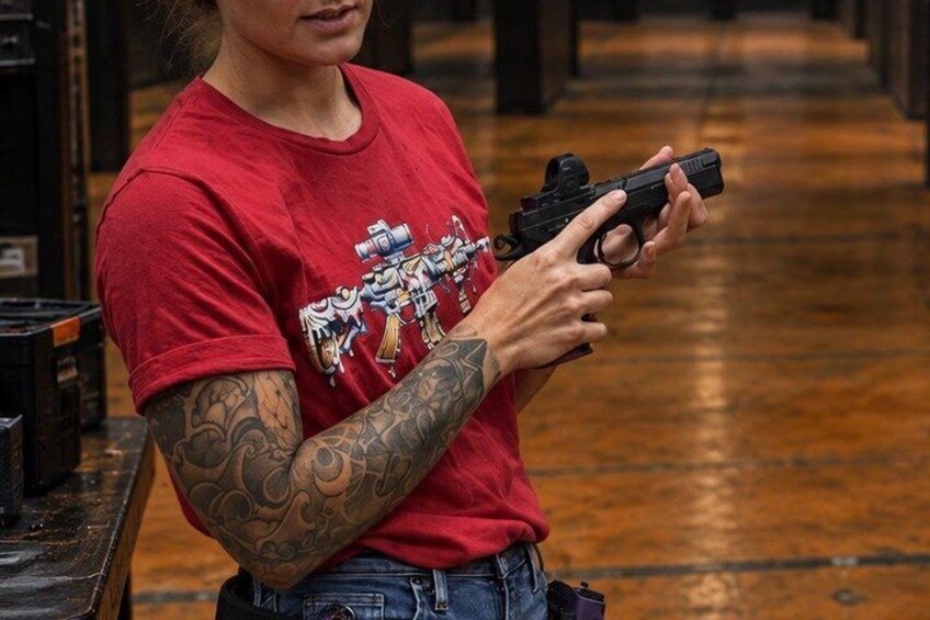 A person holding a handgun, wearing a red t-shirt with a graphic design, and having tattoos on their arms. 4