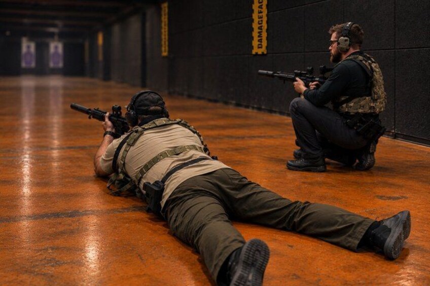 A person lying prone on the floor aiming a rifle in a shooting range with another person kneeling in the background. 4