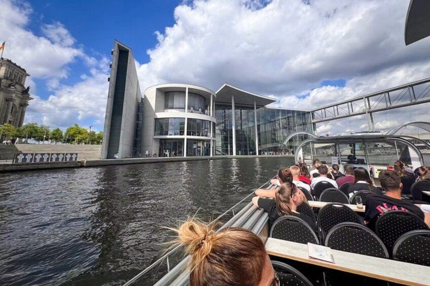 A boat tour with passengers, a modern building with glass facades, and a body of water. 3