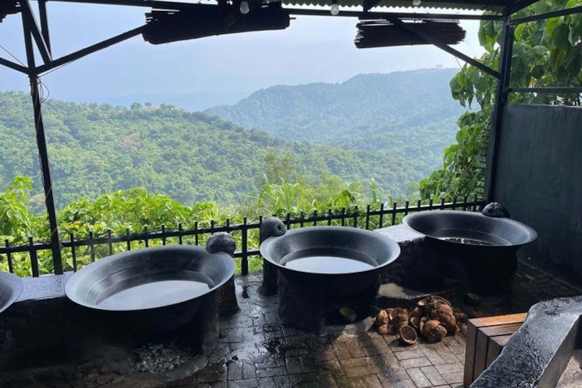 A rustic outdoor kitchen with large cooking pots and a wooden table, overlooking a lush green valley. 3