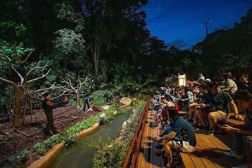 A wooden deck with people sitting and walking along a riverbank, surrounded by trees and greenery. 3