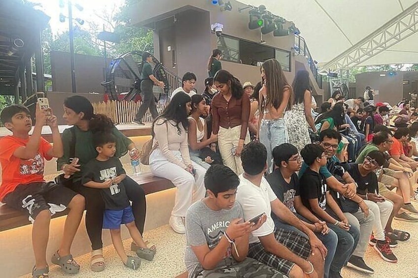A crowd of people seated on benches, some holding phones, under a covered area with a stage in the background. 5