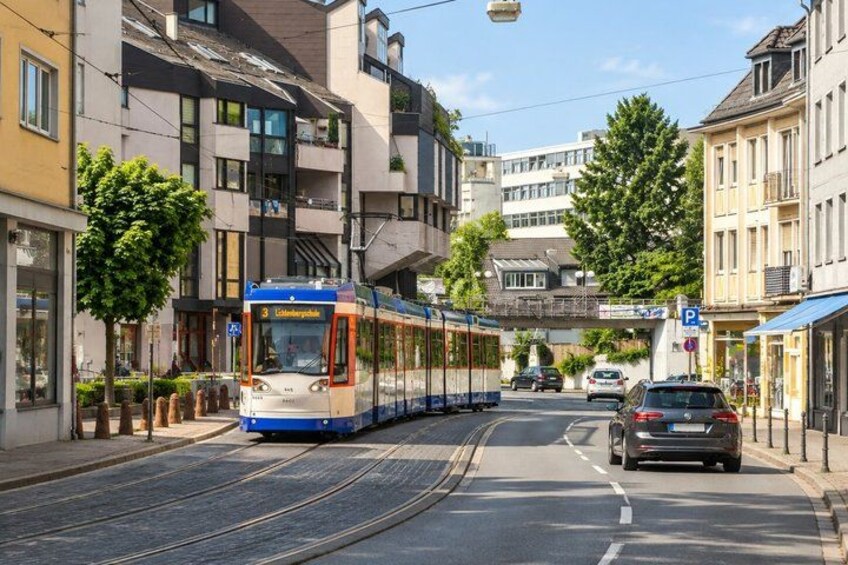 A tram on a city street with buildings and a pedestrian bridge in the background. 2