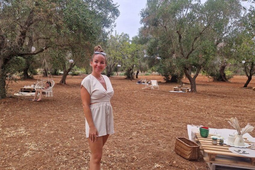A woman in a white dress standing in an orchard with trees and outdoor furniture. 4