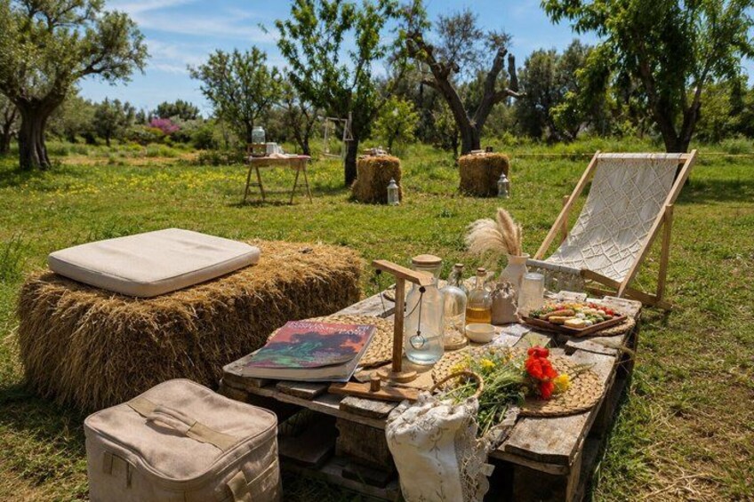 An outdoor picnic setup with a table, hay bales, a blanket, a chair, and various food items. 4