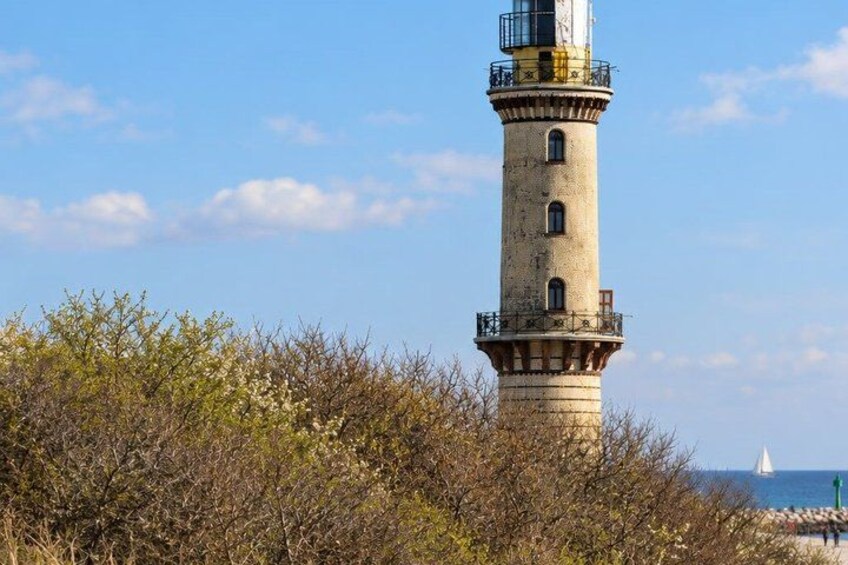 A lighthouse with a clear sky, a sailboat in the distance, and dense shrubbery in the foreground. 3