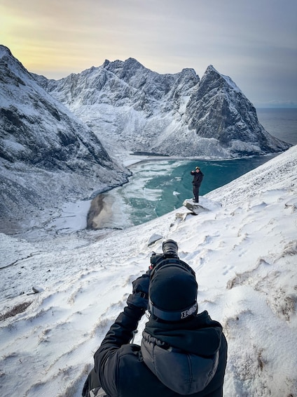 A person in a snowy mountainous landscape aiming a camera at a distant figure standing on a rock. 4