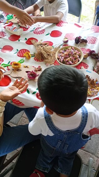 Niño sentado a una mesa con mantel estampado en rojo y blanco, rodeado de diversos materiales de manualidades y un cuenco de flores secas. 3