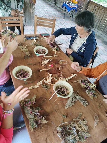 Un grupo de personas participan en una actividad artesanal, posiblemente creando coronas, con diversos materiales como flores y ramas extendidos sobre una mesa de madera. 1