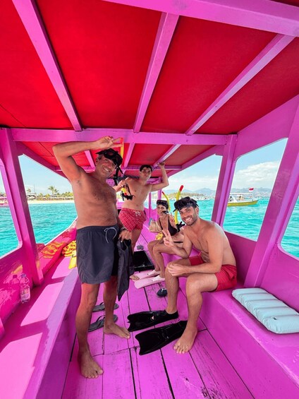 Un grupo de personas en un barco con un interior rosa brillante, algunas usando trajes de baño y snorkels, con vista despejada al océano y a islas distantes. 3