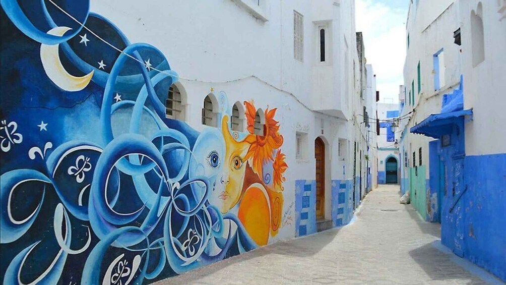 Eine enge Straße mit farbenfrohen Wandmalereien, darunter abstrakte Designs und eine große, sonnenähnliche Figur. 1