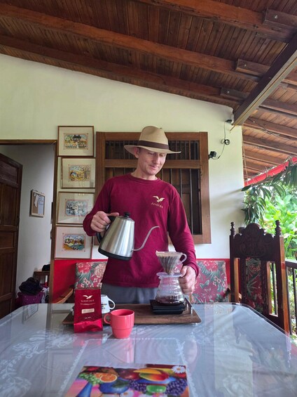 A person pouring coffee from a kettle into a coffee pot on a table with a red mug and a coffee bag. 1