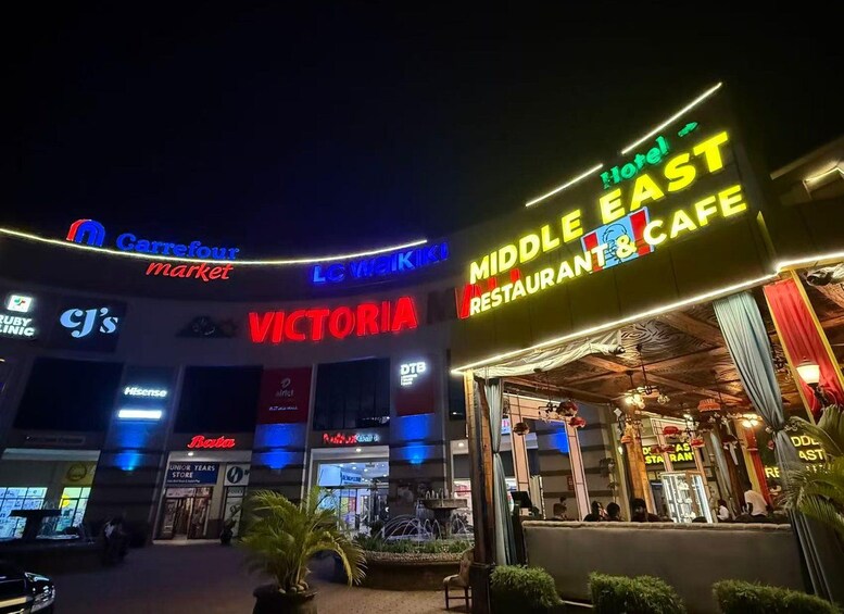 Una escena nocturna en la calle con letreros iluminados para un supermercado Carrefour, Victoria, y un café restaurante de Oriente Medio. 2
