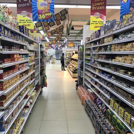 Un pasillo de supermercado con estantes llenos de diversos productos, pancartas promocionales colgando del techo y una persona comprando al fondo. 4