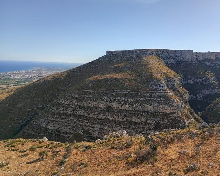 Formación rocosa en capas con cima plana, rodeada de vegetación escasa y una vista distante de una ciudad costera. 1