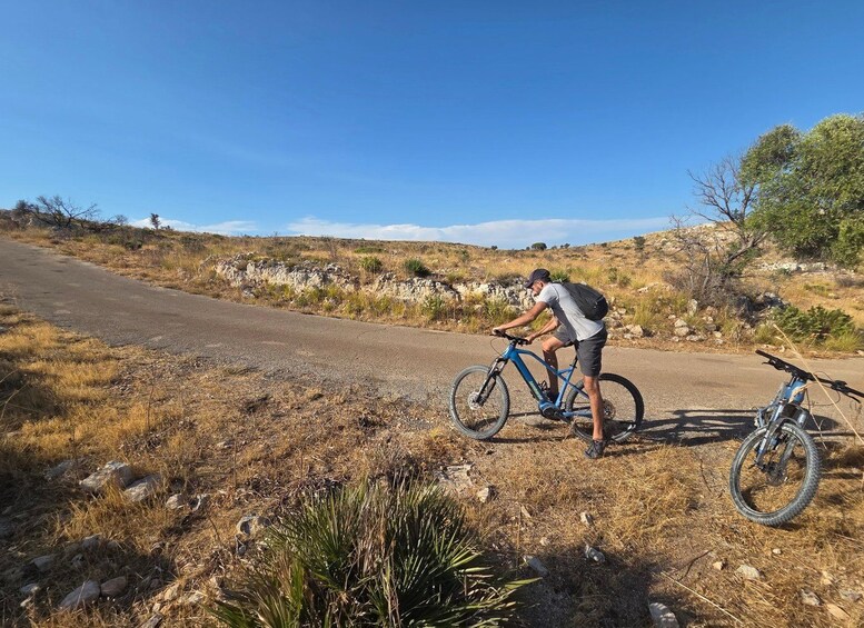 Una persona montando una bicicleta de montaña azul por un camino de tierra con vegetación escasa y terreno rocoso. 2