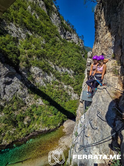 Two people sitting on a rock ledge with a rope and harness, surrounded by greenery and a clear blue sky. 3