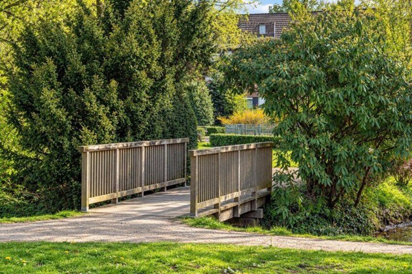 A wooden bridge over a gravel path, surrounded by dense greenery and a house in the background. 5