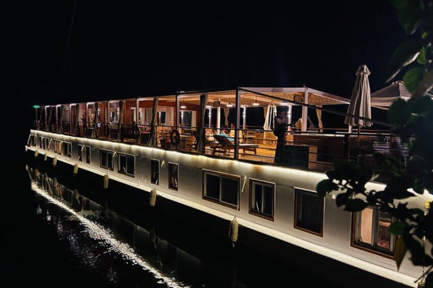 A riverboat with illuminated windows and a deck area, floating on calm water. 1