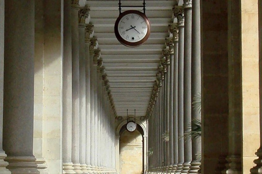 A clock hanging from a ceiling in a corridor with columns. 2