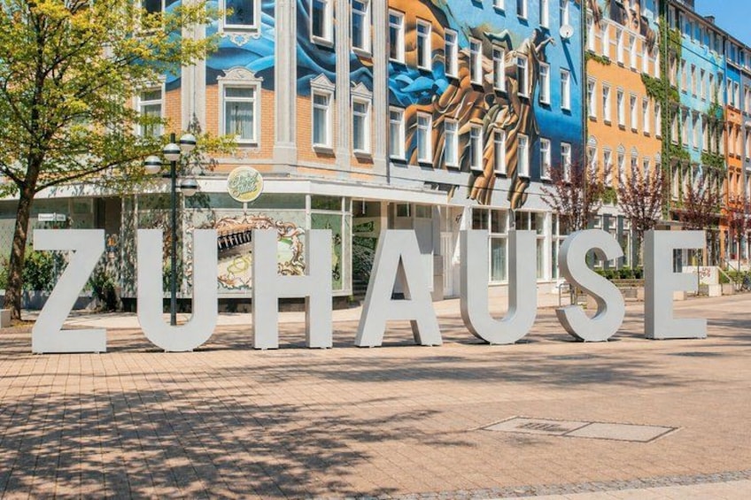 A large sign with the word 'ZUHAUSE' in white letters, set against a backdrop of colorful buildings and a tree. 2