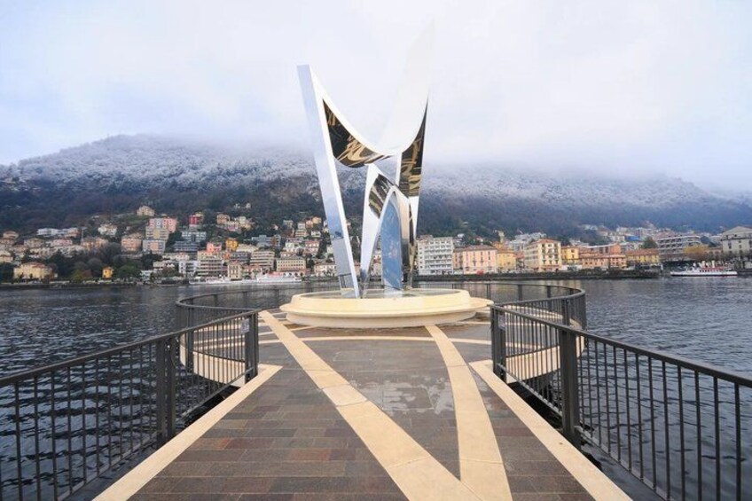 A waterfront promenade with a modern sculpture, a river, and a mountainous backdrop. 3