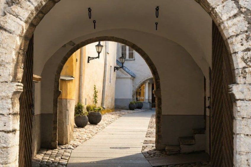 An arched stone pathway leading through a building with a tiled floor, stone bollards, and a wooden door. 2