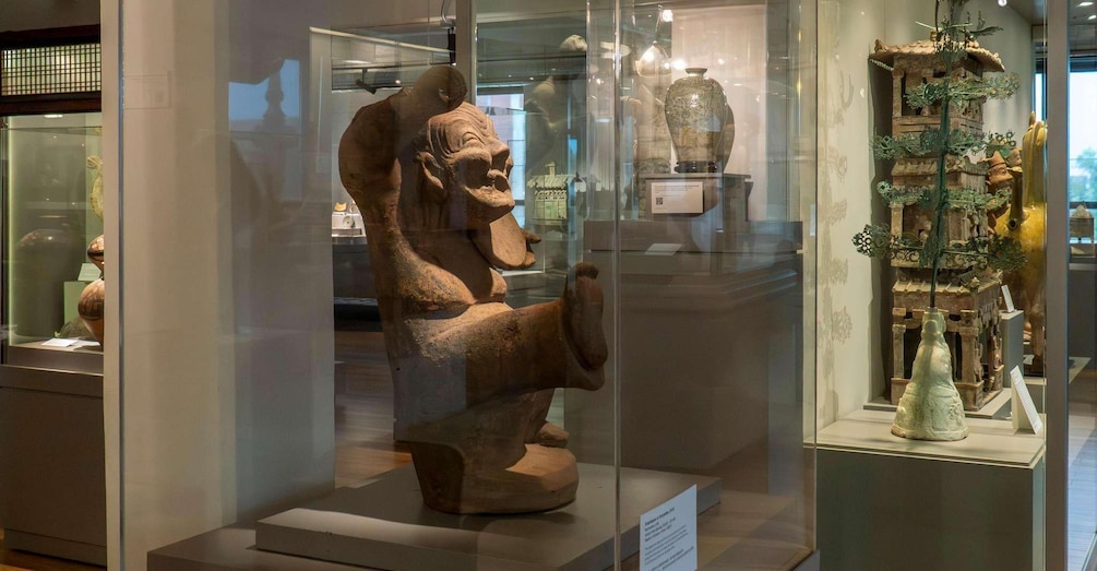 A museum display with a large ancient sculpture of a seated figure, a glass case with a smaller statue, and a decorative tree structure. 3