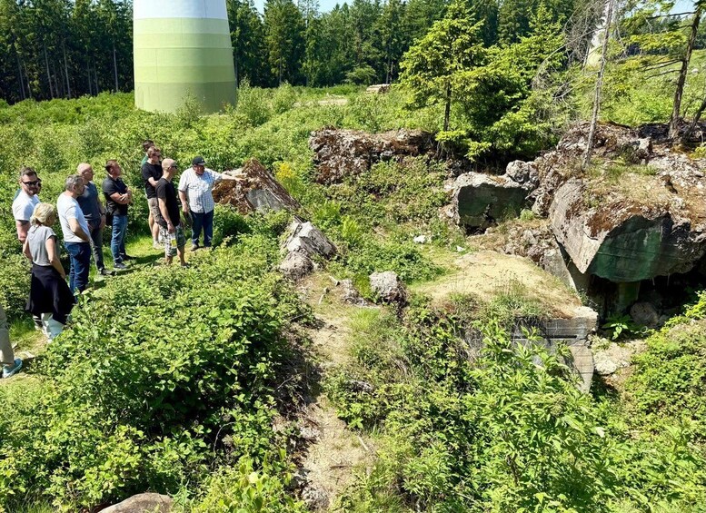A group of people standing near a large rock formation in a forested area with a water tower in the background. 3