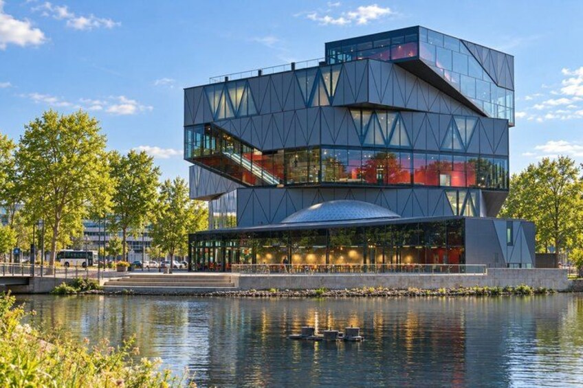 A modern building with a glass facade and a geometric design, situated by a calm body of water with trees in the background. 3