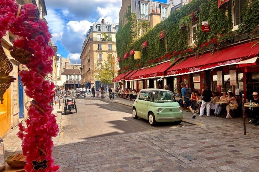 A street with a green car, red awnings, and people sitting at outdoor tables. 1