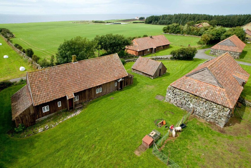 En landejendom i landlige omgivelser med flere træbygninger og en stengrunding, omgivet af grønne marker og et vandareal. 5