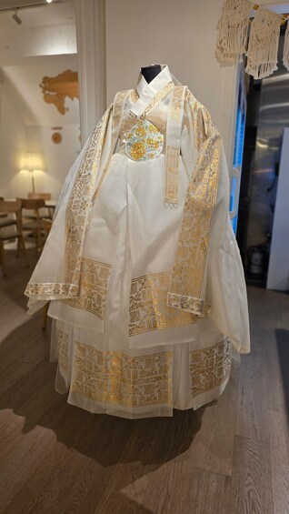 Eine Schaufensterpuppe trägt ein traditionelles, weißes Gewand mit Goldstickereien und steht in einem Raum mit Holzboden und einer Wanddekoration aus Makramee. 5