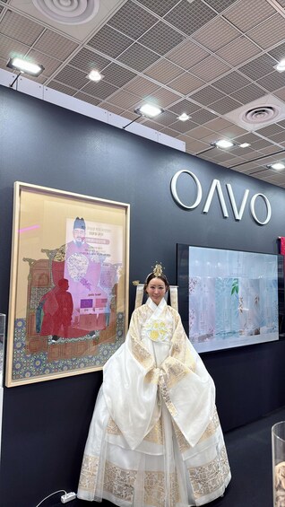 Eine Person in traditioneller Kleidung steht vor einer Wand mit dem Schriftzug 'OAVO' und gerahmten Kunstwerken. 1