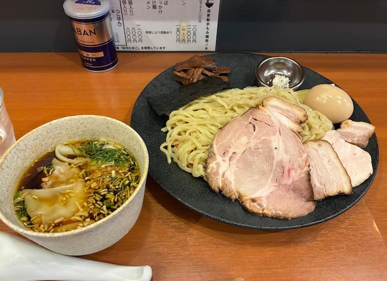 A bowl of ramen with noodles, pork, and vegetables, a plate with sliced pork and a boiled egg, and a container of sauce. 5