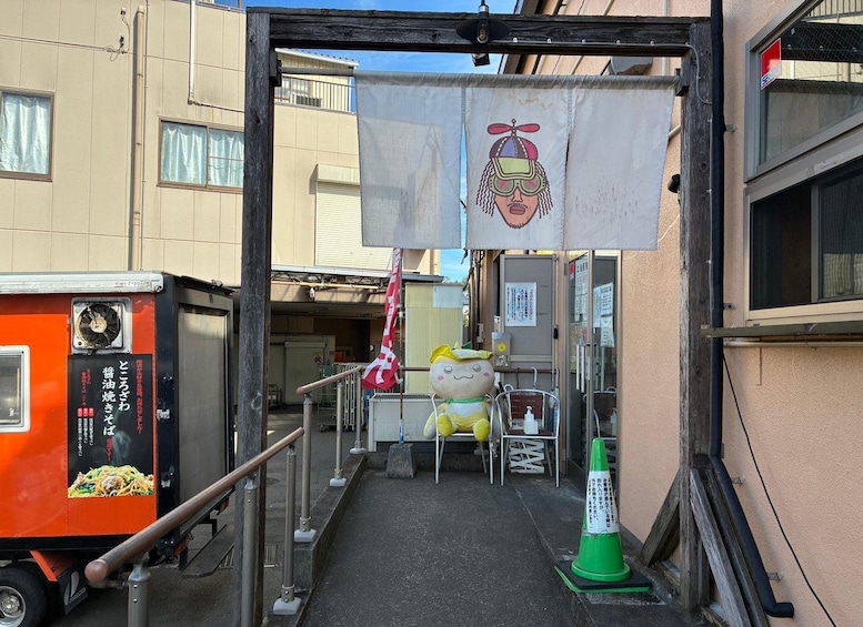 A narrow alleyway with a small shop entrance, a red and white flag, a green traffic cone, and a cartoon character decoration. 2