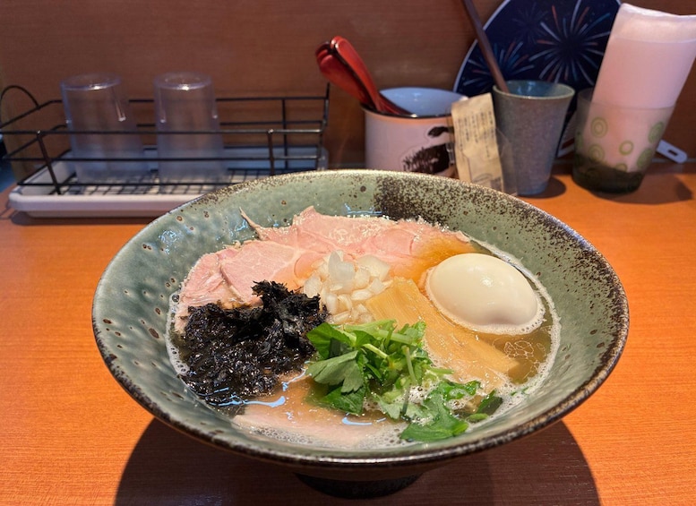 A bowl of ramen with pork, egg, seaweed, and greens on a wooden table. 1
