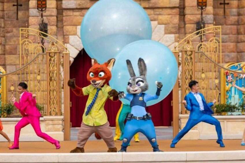A person in a fox costume, a person in a rabbit costume, and a person in a blue suit dancing in front of a building with large blue balloons. 2