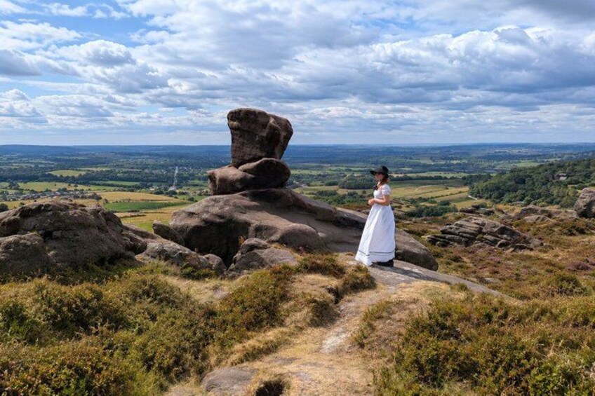 A person in a white dress standing on a rocky outcrop with a view of a valley and distant hills. 4