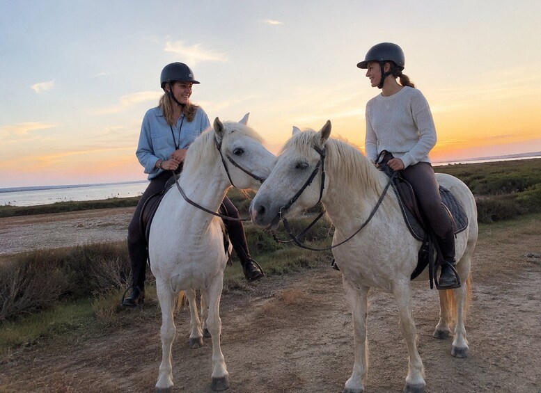 Dos personas montando caballos blancos por un sendero de tierra, con un atardecer de fondo. 2