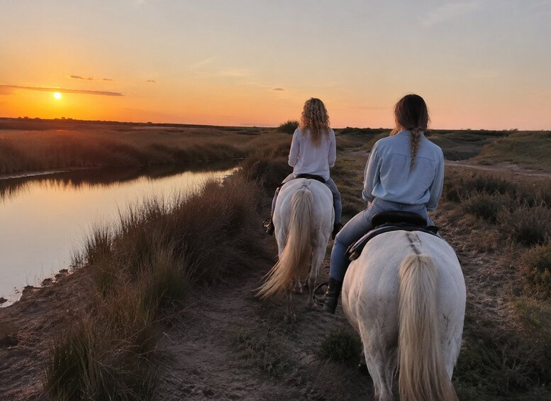 Dos personas cabalgando a caballo por un sendero de tierra junto a un cuerpo de agua al atardecer. 4