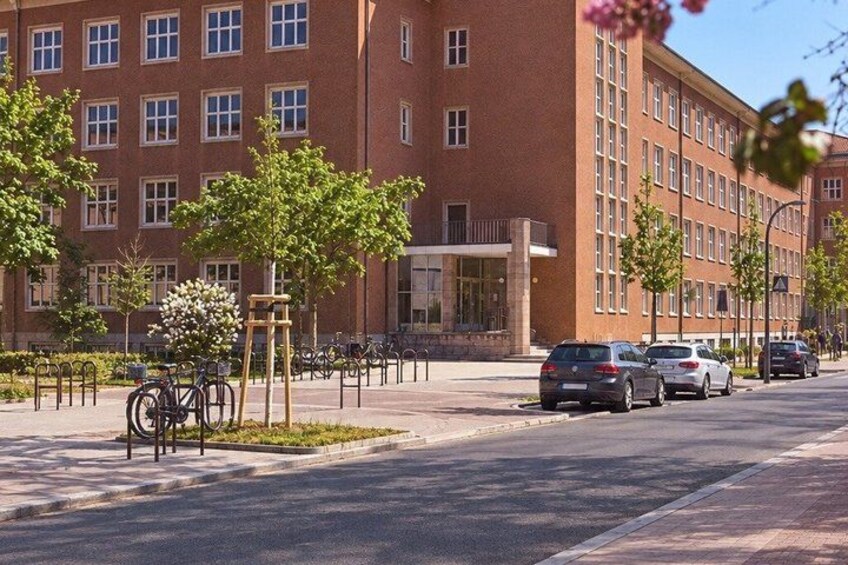 A red brick building with multiple windows, a sidewalk with trees, parked cars, and bicycles. 4