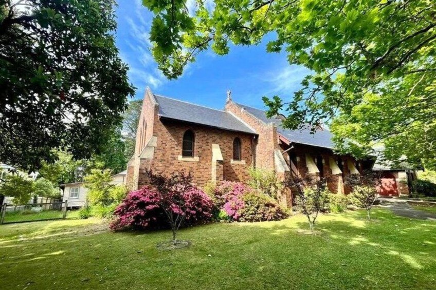 A brick church with a steeple, surrounded by green grass and flowering bushes. 5