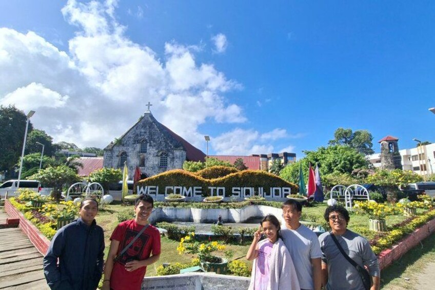 A group of people in front of a sign that reads 'WELCOME TO SIJOURO' with a church and a flag in the background. 1