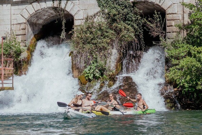 A group of people rafting on a river with a waterfall and stone arches in the background. 1
