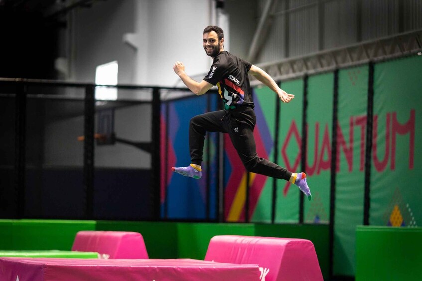 A person performing a high jump over a pink obstacle in an indoor sports facility with a green wall and the word 'QUANTUM' visible. 1