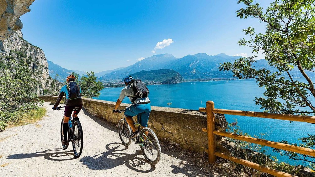 Dos ciclistas en un sendero de grava con baranda de madera, con vista a un lago y montañas. 3
